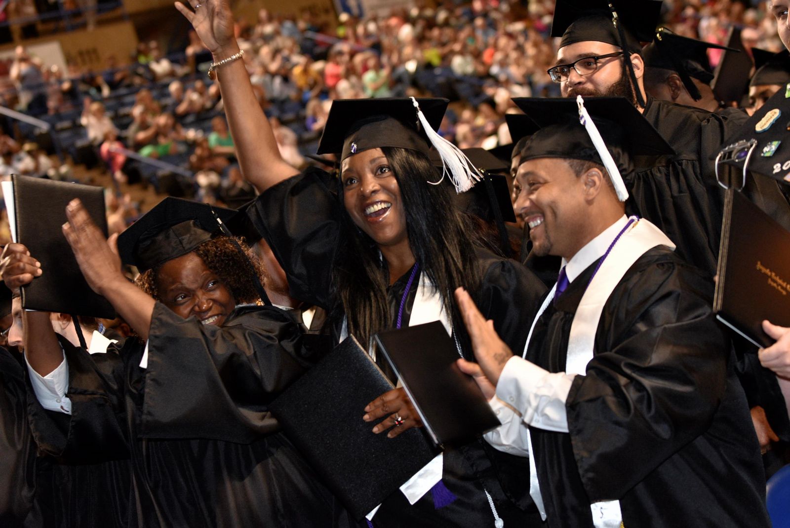 A forest green, hyperlinked, button labeled Technical Certificate of Credit in white, bolded, underlined text with an image of a crowded gathering of graduates and family focusing on four graduates wearing black graduate caps and gowns with white tassels and stoles: an African American female smiles while holding a black diploma/degree holder up with her left hand placed on the side, to her left an African American female smiles and raises her right hand wearing a metal bracelet while holding her black degree/diploma holder in her left hand, an African American male stand smiling and holding his black diploma/degree holder in his right hand with his left hand held up behind it, and a Caucasian male with a  black beard and glasses stands behind and to the right of those three.