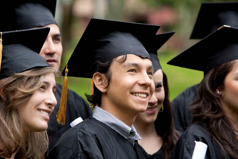 A forest green, hyperlinked, button labeled Associate Degrees in white, bolded, underlined text with an image of six graduates smile to the right of the frame in black graduate caps and gowns: A Caucasian female with sandy, brown hair stands to the right of a smiling, brown haired, Caucasian male with a blue collared shirt showing above his gown. To his left a brown haired, Caucasian female stands smiling with another brown haired, Caucasian female standing partially out of the frame to her left. A Caucasian male stands partially blocked by the first female and a sixth graduate stands hidden behind the last female.