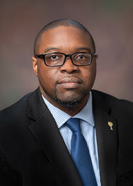 Dr. Jermain Whirl, an African American male, wears a black suit jacket with a gold pin, a blue and white striped, collared shirt; a cerulean blue tie and black square glasses. He is pictured against a foggy brown background.