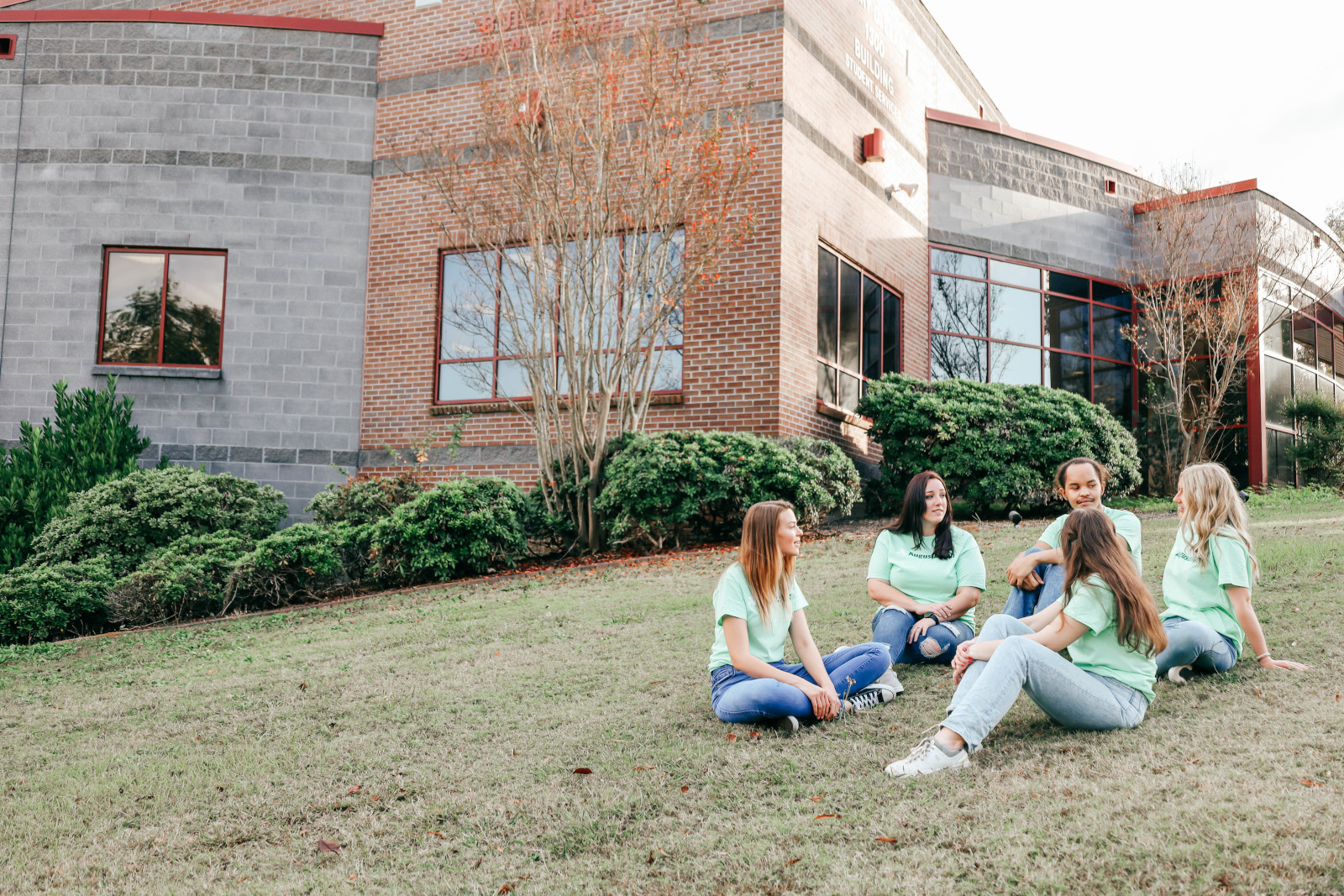 A group of students all wearing mint green Augusta Technical College shirts sit on the lawn to the left of the 1300 bldg chatting.