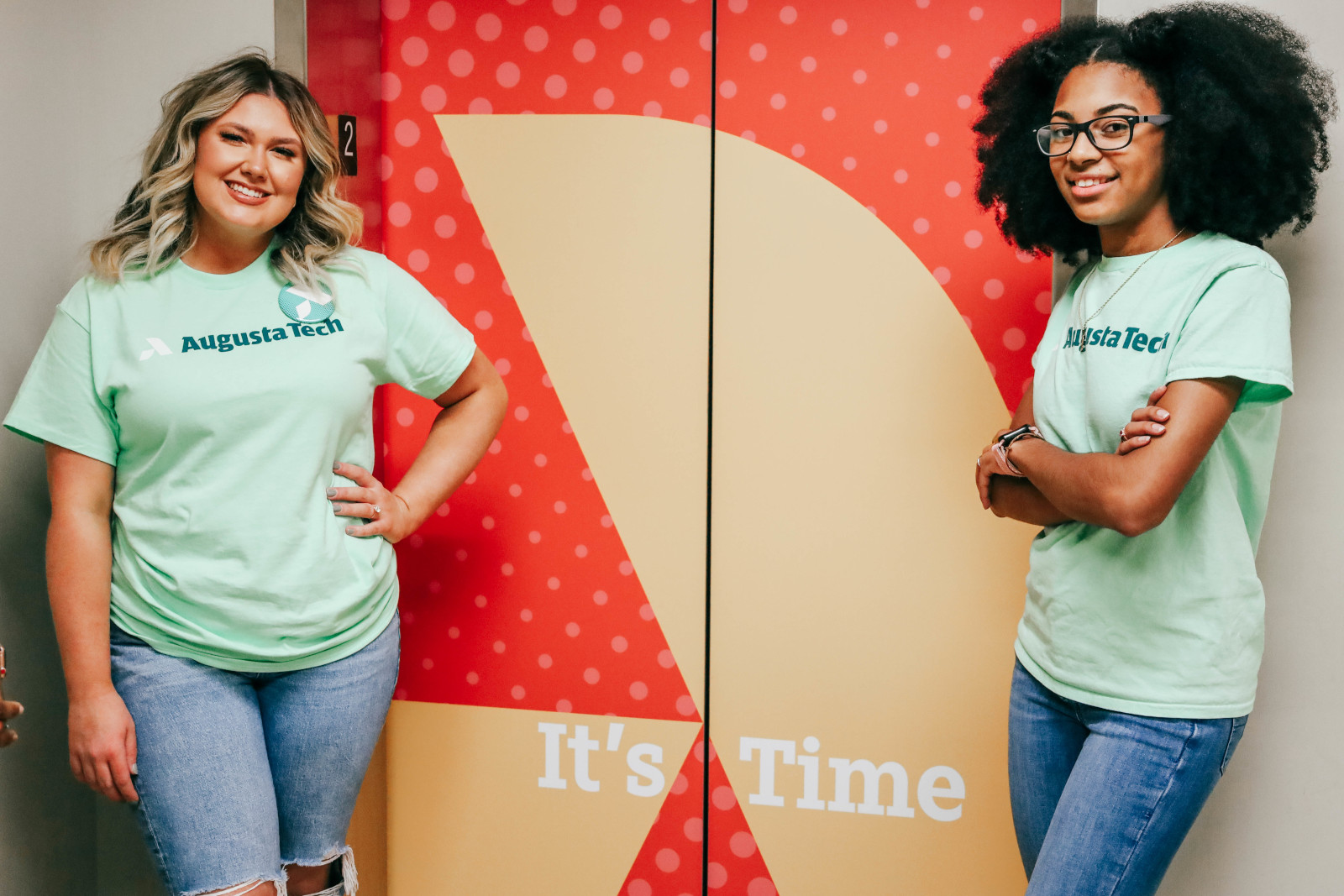 Two female students one a Cacasian female with long blonde hair standing on the left and the other an African American female with shoulder length natural hair on the right, stand wearing mint green Augusta Technical College shirts in front of the 1400 bldg elevator marked by an orange background with a yellow Augusta Technical College A icon and the motto It's Time.