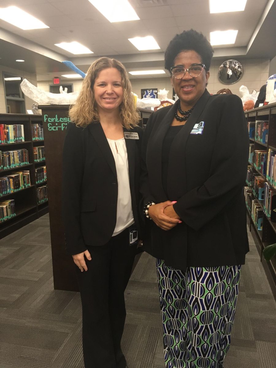 A blonde, caucasian female, Dr. Holly Barnes(Principal) wearing a black jacket with a white shirt and black dress pants stands with an African American female, Bonita Jenkins(Augusta Technical College), wearing a black jacket with a black shirt and a black necklace pictured with a library backdrop.