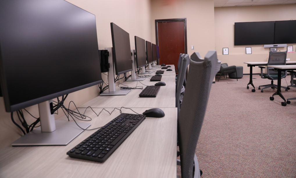 A picture showing the length of a table holding four computers against a wall. Two flat screen tvs are on the back wall with rows of tables and chairs in the center of the room.