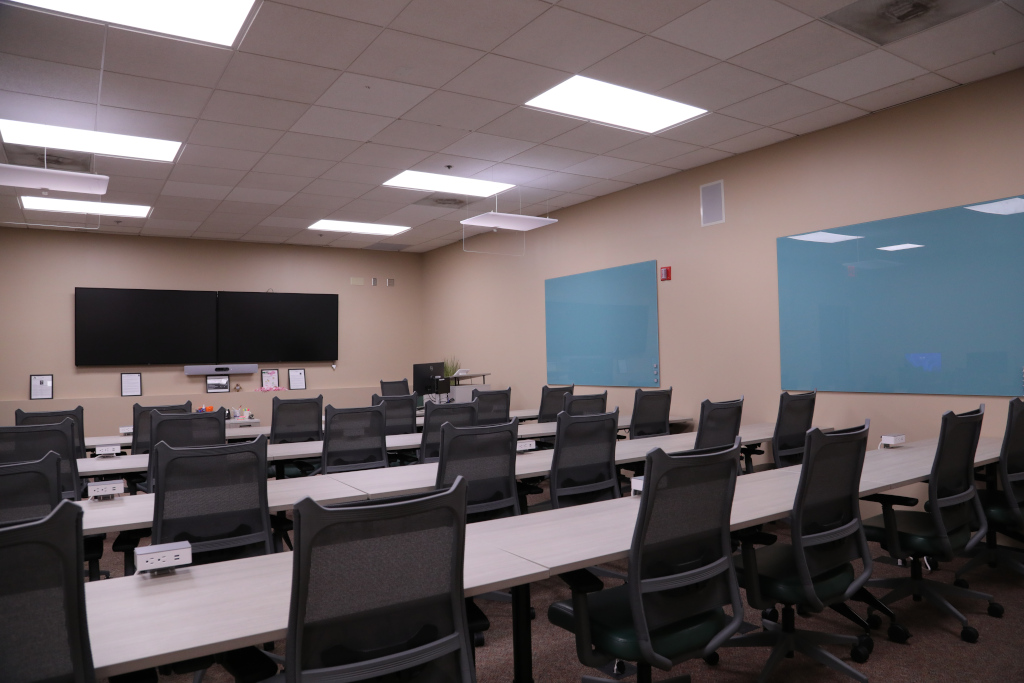 Two TVs are mounted on the far wall with rows of tables and chairs facing them. Two blue squares are on the right wall.