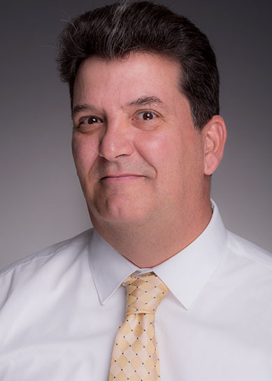 A Caucasian male with brown hair, Patrick Riley wears a white collared dress shirt and a gold diamond pattern tie with the diamond points separated by single black dots.