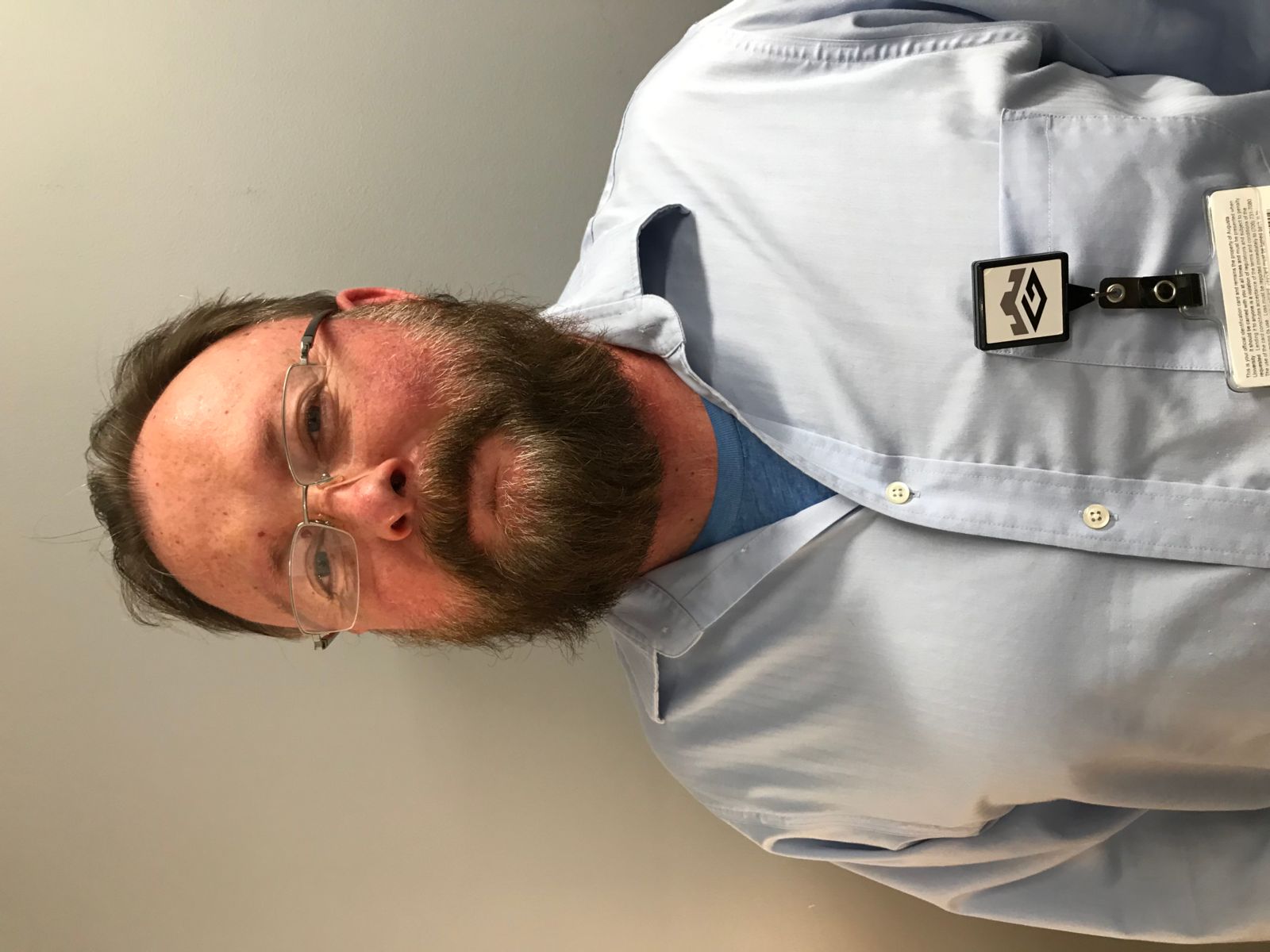 A Caucasian male with brown hair and a beard, Chester smith has rimless glasses and is wearing a blue tshirt under a light blue, collared, button down dress shirt.