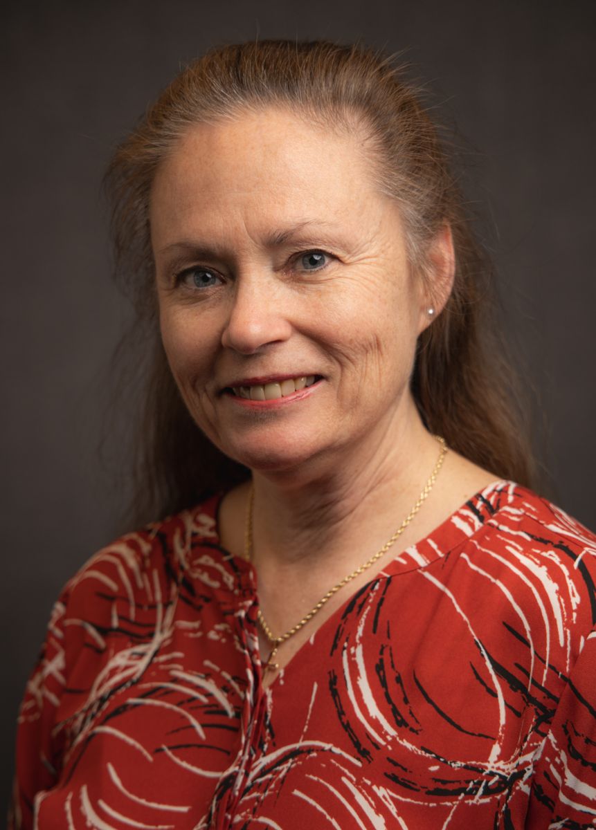 A Caucasian female with long brown hair, Gail Volz smiles at the camera and is wearing a red top with a black and white paint circle pattern on it as well as a chain necklace.