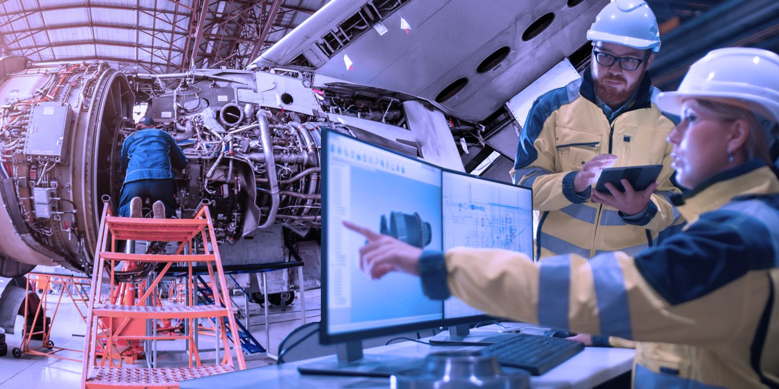 A mechanic works on an airplane engine while kneeling on an orange safety ladder. A male and a female engineer wearing yellow and black jackets and hardhats study plans on a computer monitor and tablet.