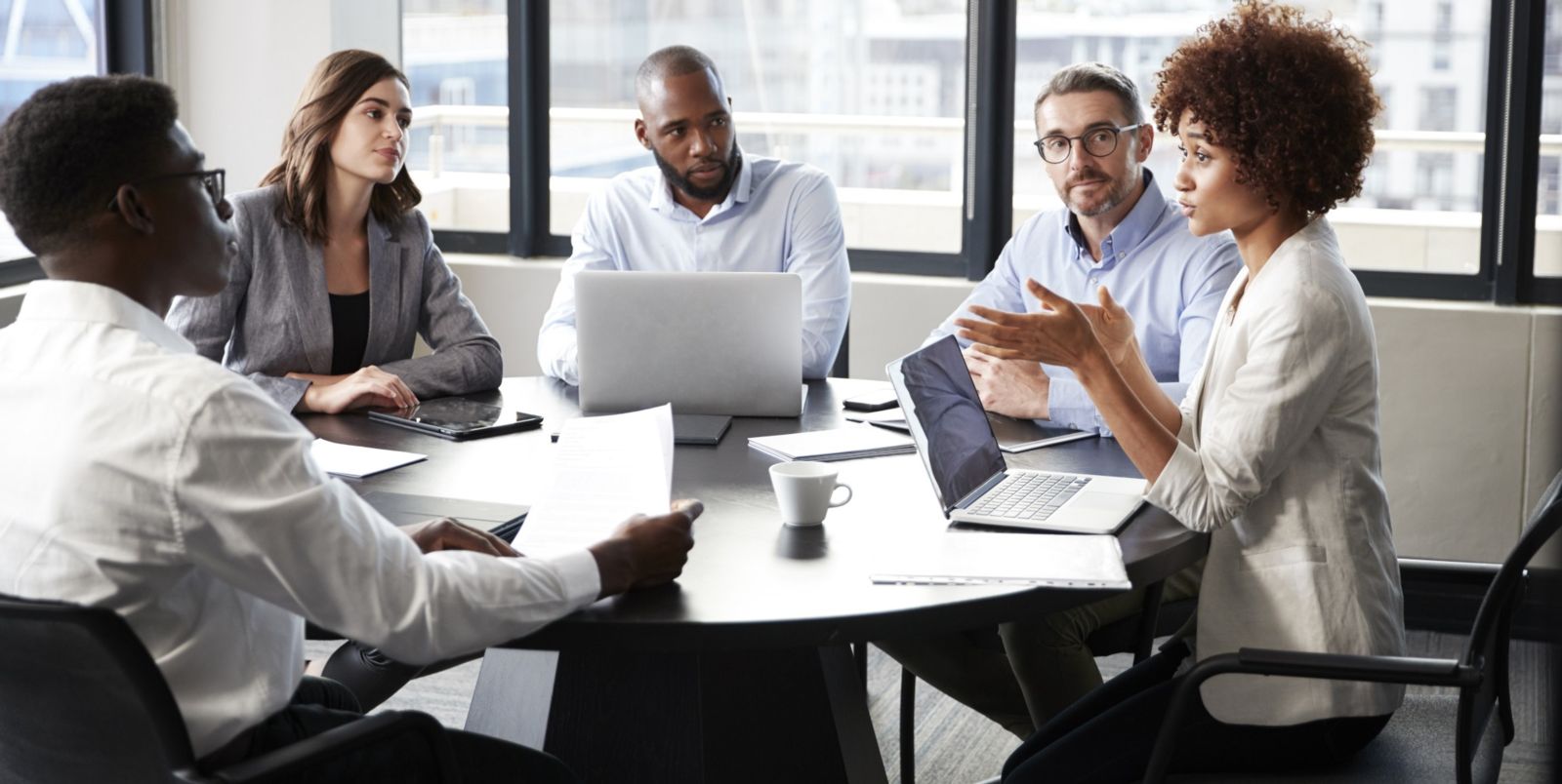 Five individuals composed of two African American males, one African American female, a Caucasian male, and a Caucasian female wear business casual clothes and sit around a round table discussing business using computers and paperwork.