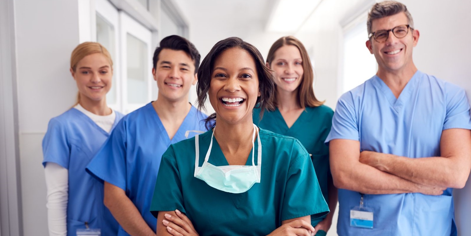 Five health care workers stand in a hallway wearing scrubs in various shades of blue. An African American female stands center with a Caucasian male and female standing directly behind her. Another Caucasian female stands to the far left behind the Caucasian male and another tall Caucasian male stands to the far right.