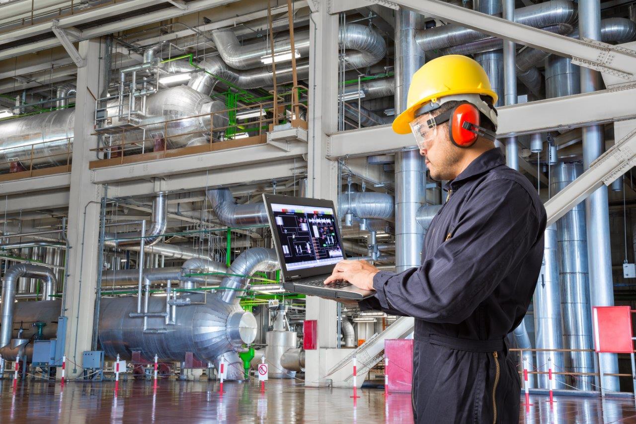 A factory worker holds a laptop while wearing a black jumpsuit, yellow hardhat, orange noise cancelling headphones, and safety glasses in a factory containing pipes and machinery.