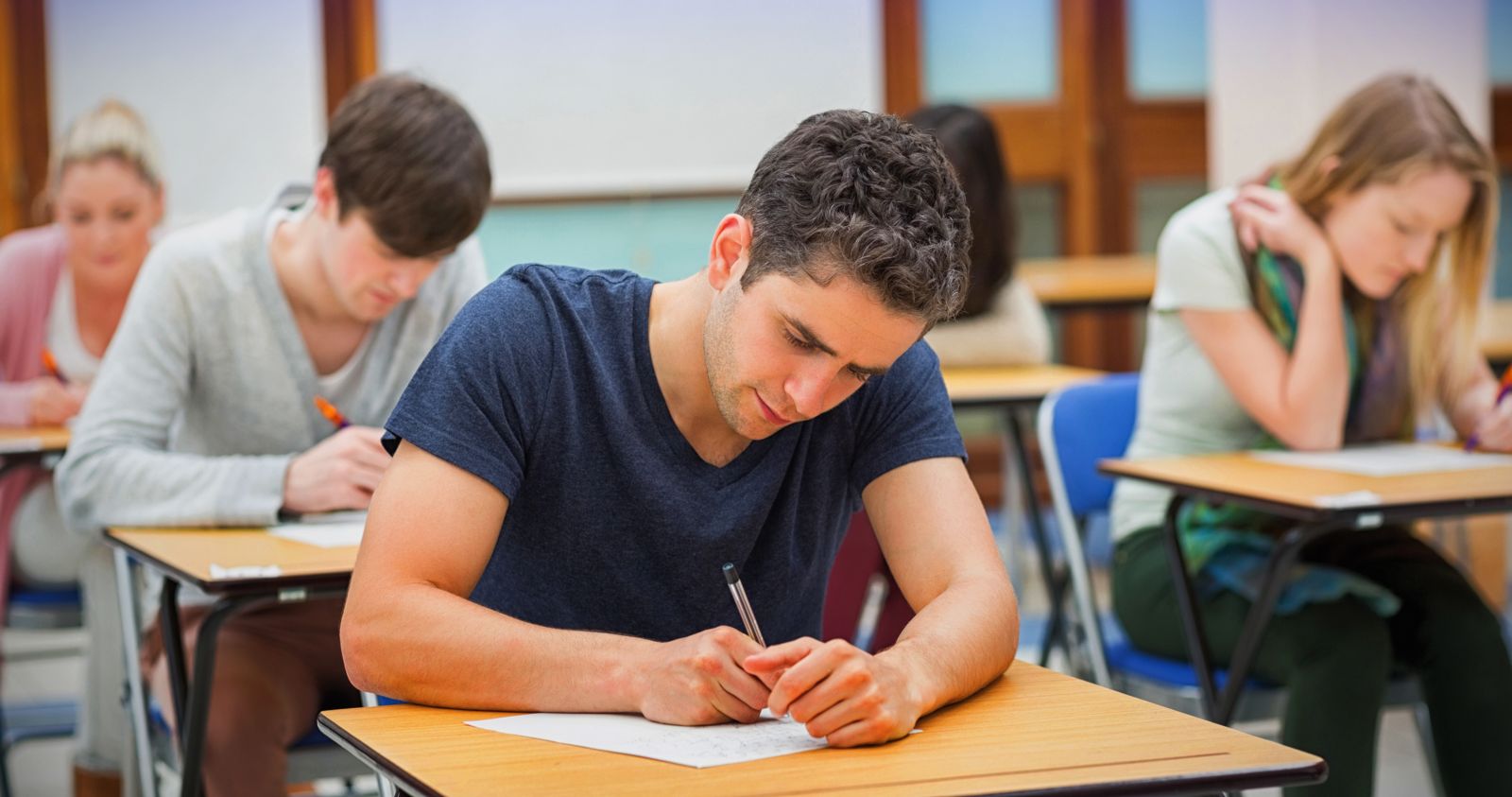 Two male and two female Caucasian students wearing casual clothing sit at desks in a classroom while writing on paper like they are taking a test.