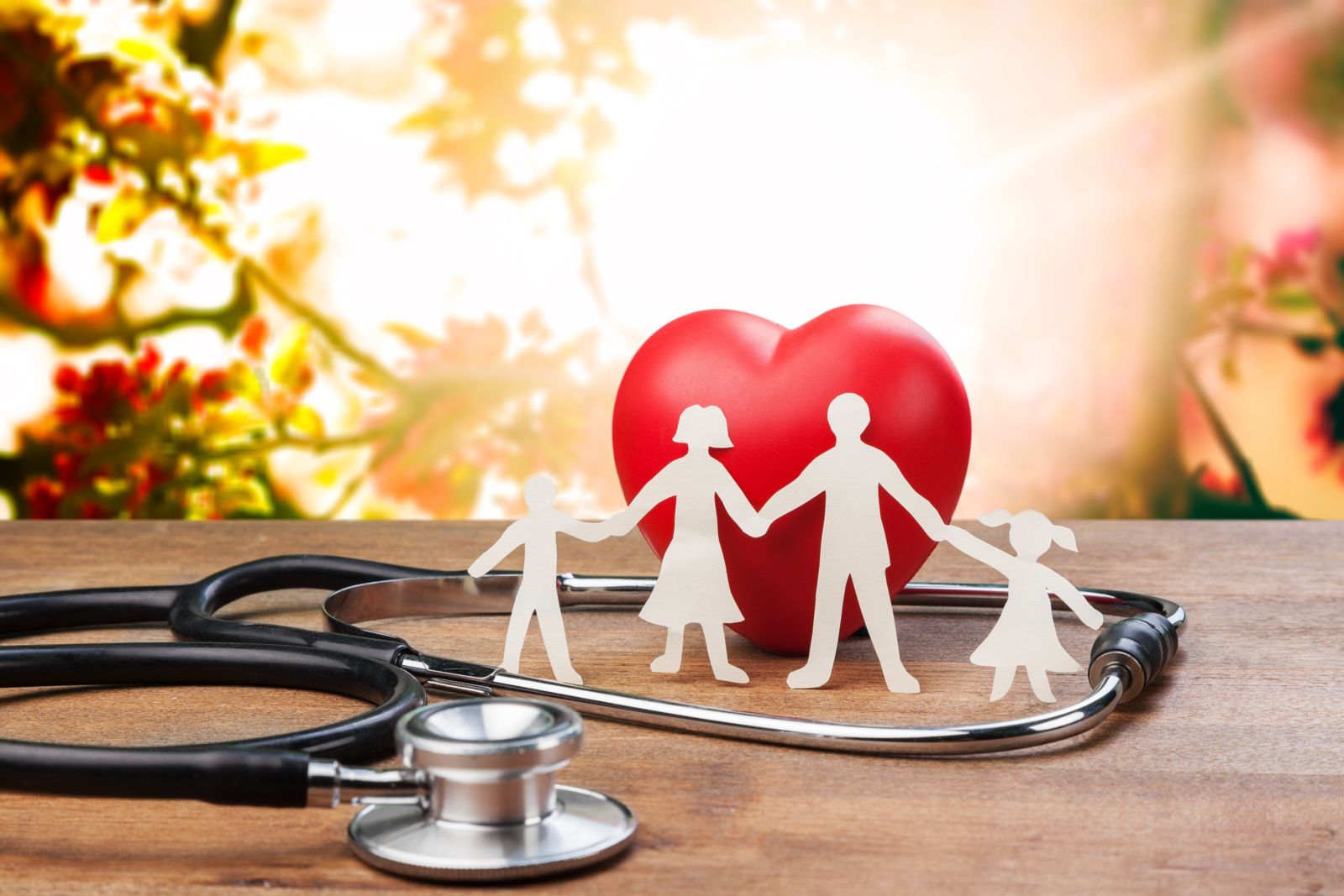 White paper cutouts of a Father, mother, and two children holding hands with a red wooden heart behind them. The heart and paper cutouts are inside the head portion of a stethoscope on top of a wooden surface with a tree branch and white light in the background.