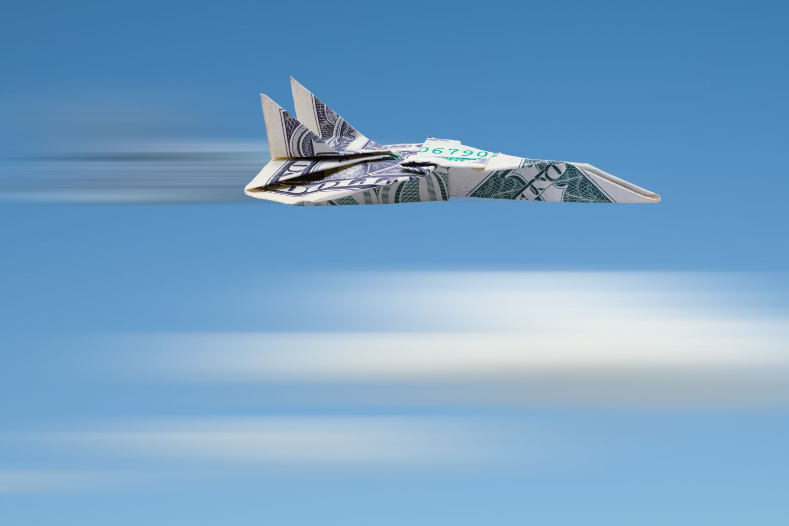 A paper airplane made from a 1-dollar bill is set against a sky-blue backdrop with white blur lines below that look like blurred clouds and black and white blur lines behind the engines to represent the plane flying at great speed.