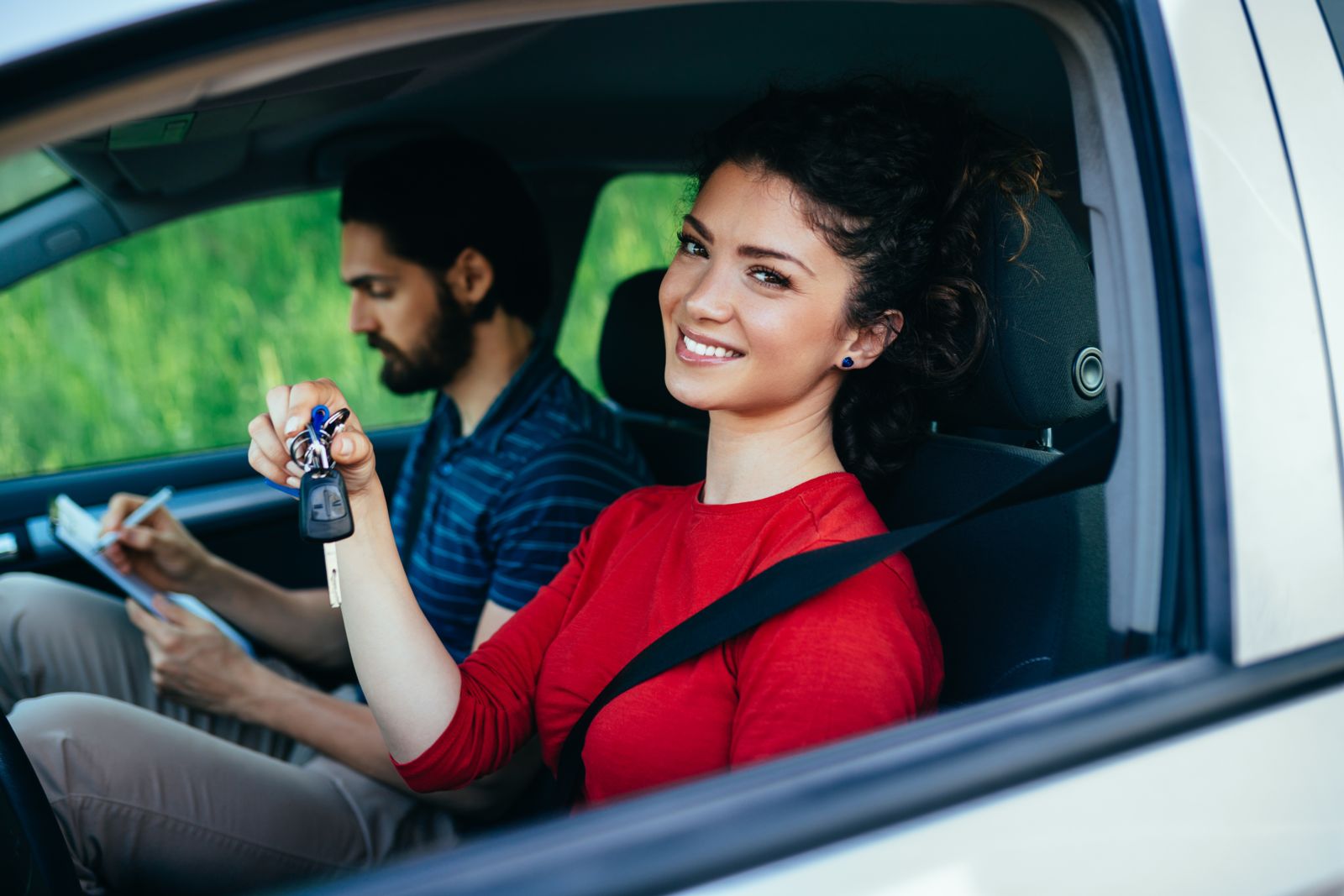 A smiling Caucasian female with black hair wearing a red long-sleeved t-shirt sits in a silver car holding up keys in her right hand while a bearded Caucasian male also with black hair and wearing business casual clothing writes on a paper using a clipboard.