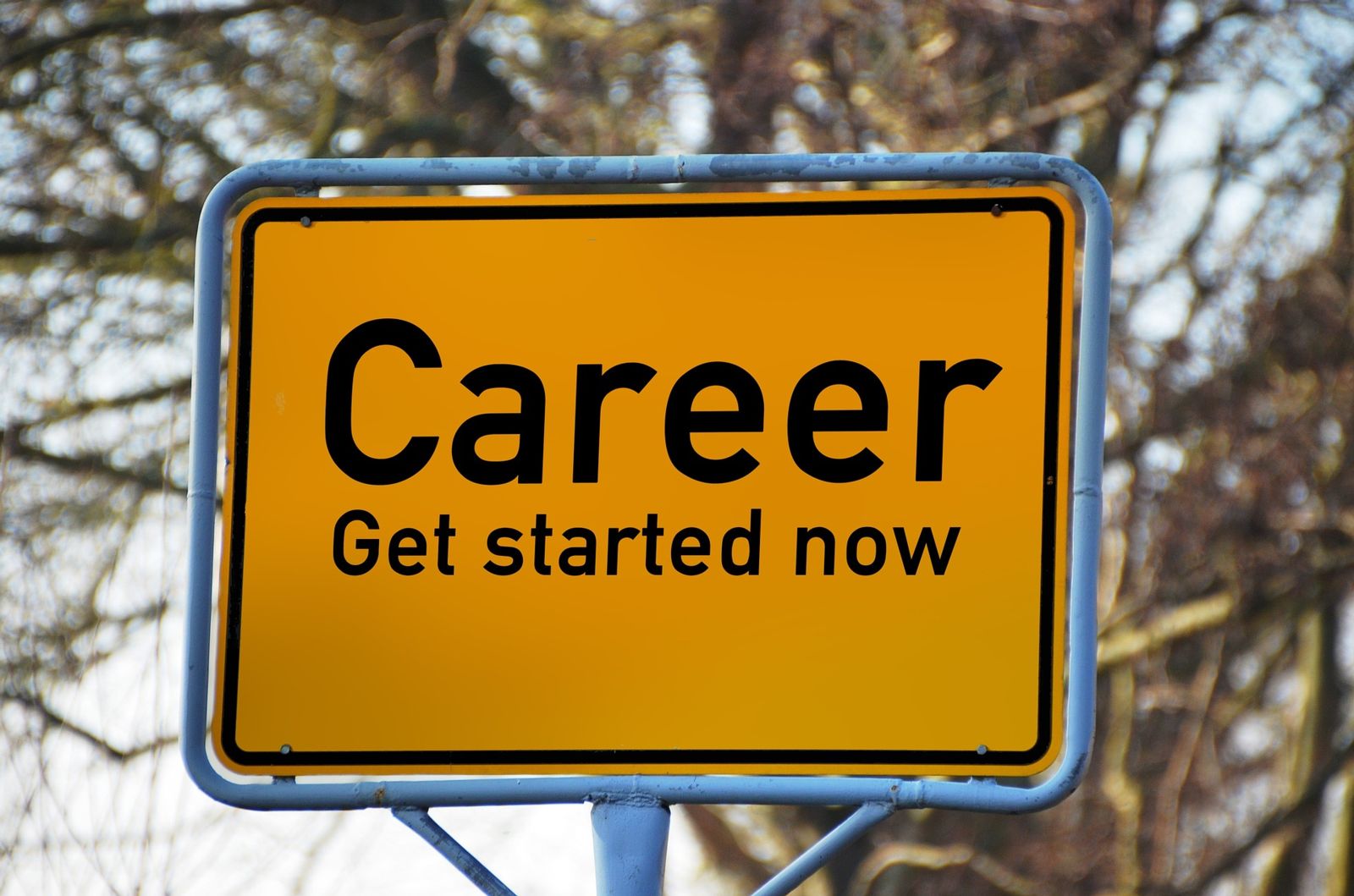 A yellow sign with the words Career Get started now in black font is displayed on a blue metal frame and pole in front of leafless trees.