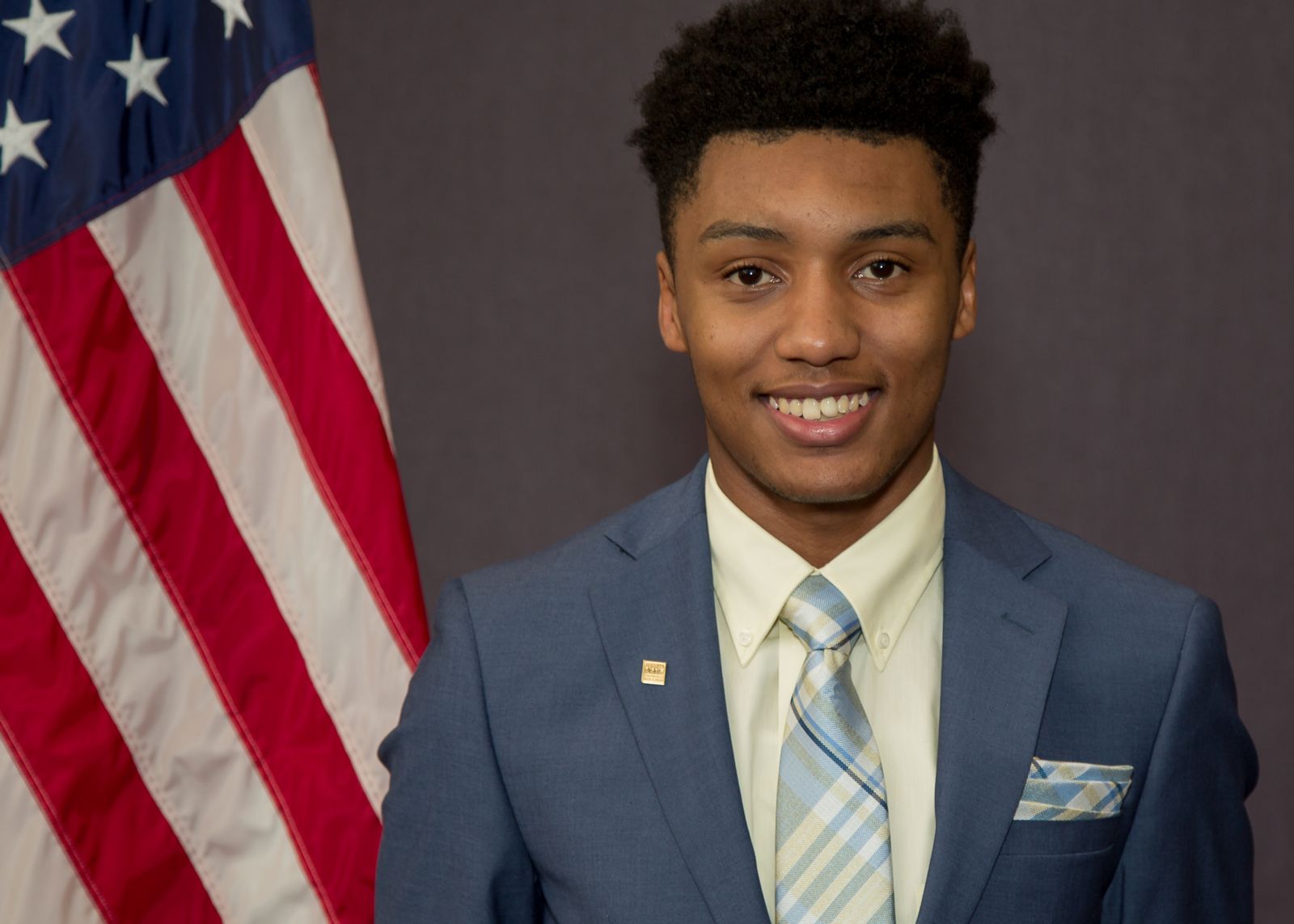 African American college student Rah'Mere Williams, dressed in a navy blue suit with a white collared shirt and light blue striped tie stands in front of and to the right of an American Flag against a grey background.