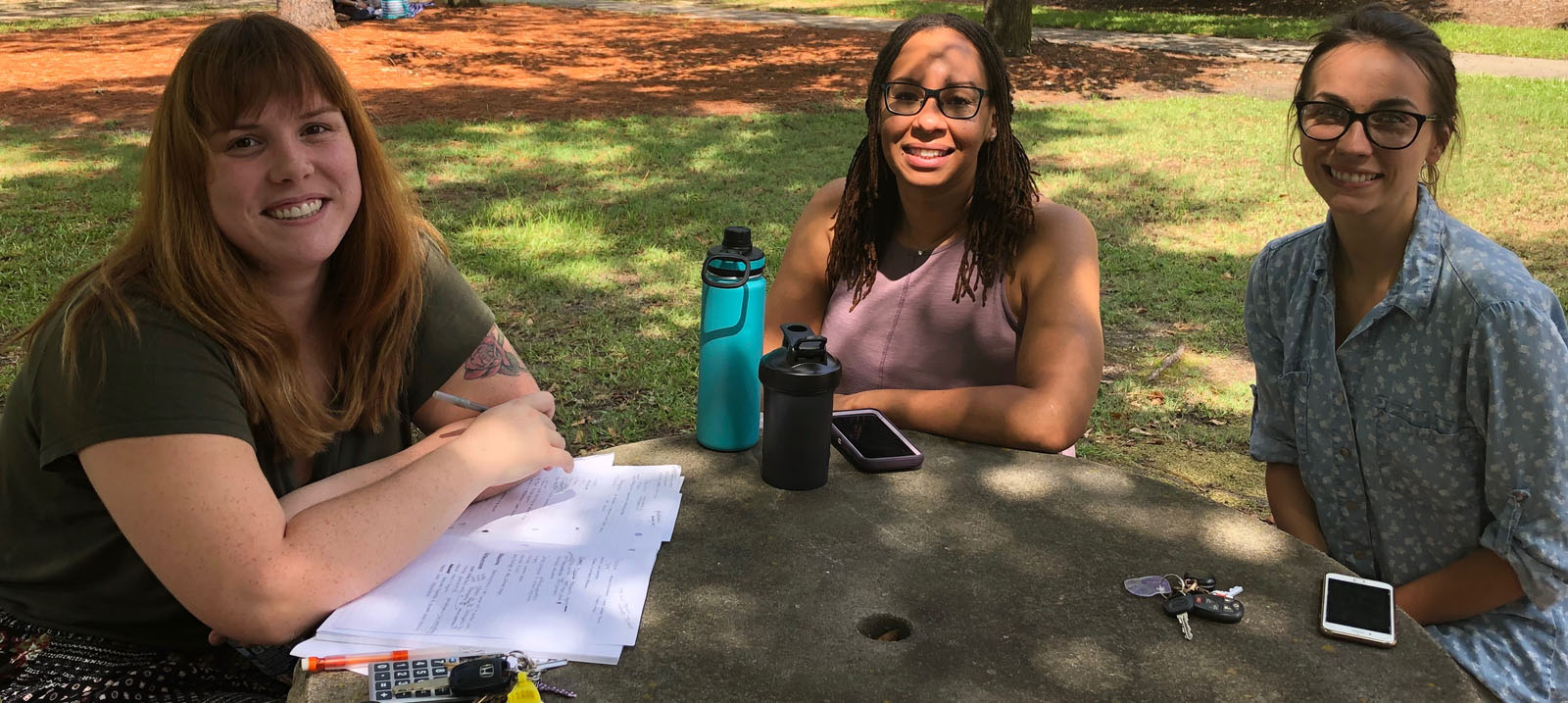 Three smiling, female, Caucasian students sitting at a round, grey stone table: the first has brown hair with blond highlights worn down and is wearing a black shirt and a black skirt with multi-colored polka dots and is pictured with a stack of paper under her crossed arms on the table with a ring of keys including yellow and white cards and a black cell phone; the second has brown hair worn down and black rimmed glasses with a lavendar pink shirt, she is pictured with a tall, teal and black water bottle, a shorter solid black water bottle, and her cell phone with a lavendar back and white edges; and the third has on a blue, collared button up shirt with white polka dots and tan pants, she is pictured with her car keys and a cell phone with white, horizontal bars on the upper and lower portion of the face.