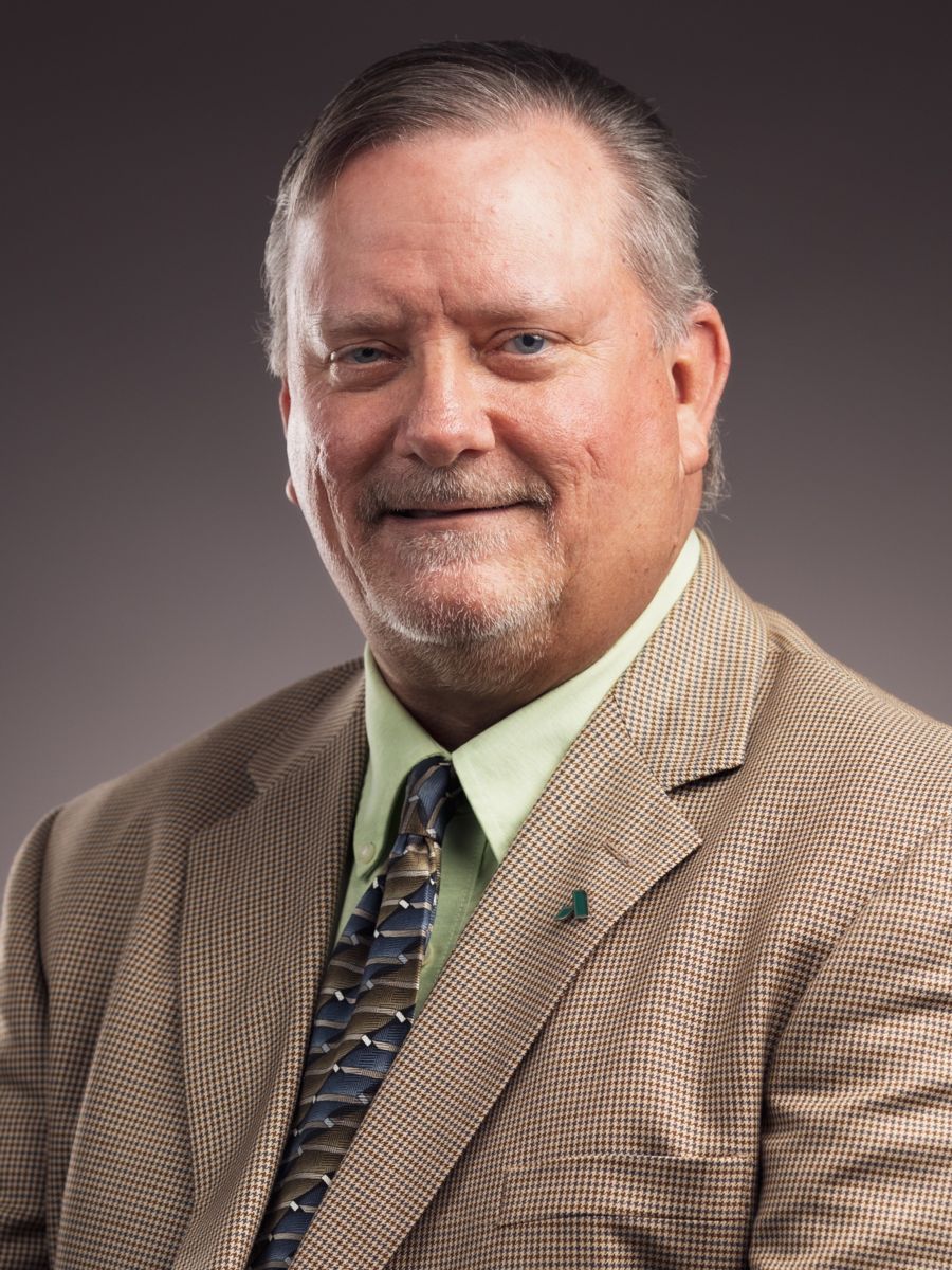Professional headshot of Mr. James Bostock wearing a tan checkered suit jacket, light green dress shirt, and patterned tie against a plain gray background.
