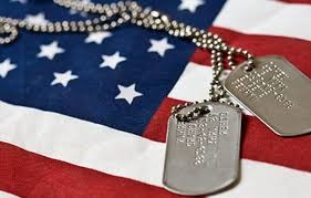 An image of the American flag showing the lower right corner of the navy blue section with eleven white stars and four red strips separated by three white strips to the bottom and right of the navy blue section. A twisted chain laying on the blue section holds a pair of dog tags laying on the stripes.