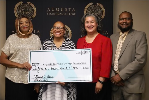 Four adults stand side by side, smiling, in front of a black backdrop repeatedly printed with the gold seal and name “Augusta Technical College.” From left to right: a woman wearing a light gold, short-sleeve, shimmery blouse; a woman in glasses wearing a black-and-white patterned dress; a woman in a red blazer over a black top; and a man wearing a tan suit with a patterned tie. The two people in the center hold a large ceremonial check made out to the Augusta Technical College Foundation in the amount of $15,000, dated 12/05/2025, designated as a scholarship. The group appears professionally dressed, and the setting suggests a formal scholarship presentation or announcement event.