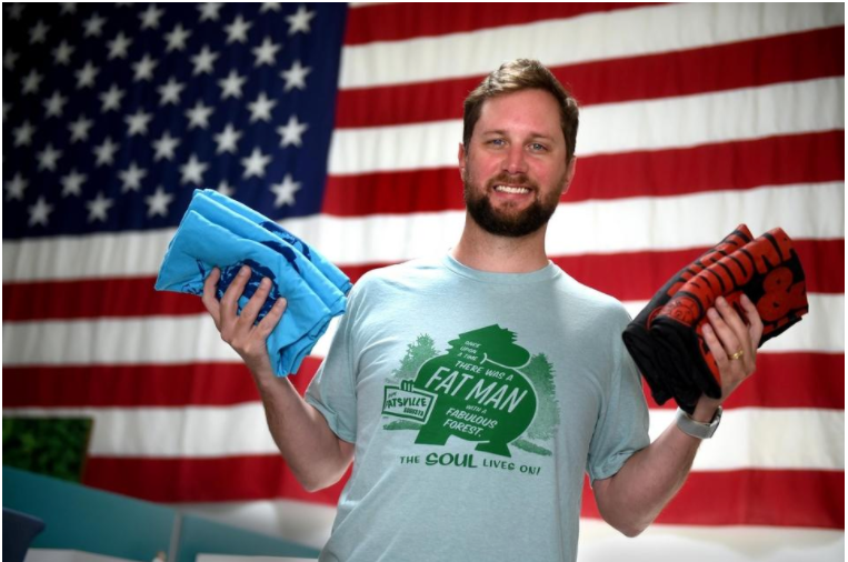 A Cacausian male with short brown hair and a beard, Sean Mooney stands in front of the United States flag smiling and holding three folded light blue t-shirts in his right hand and two folded black and red t-shirts in his left hand.