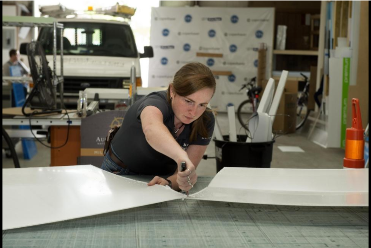 A Caucasian female with brown hair, Brittany Doster is leaning over a table while cutting a white board. A white van with design supplies and tools are in the background.