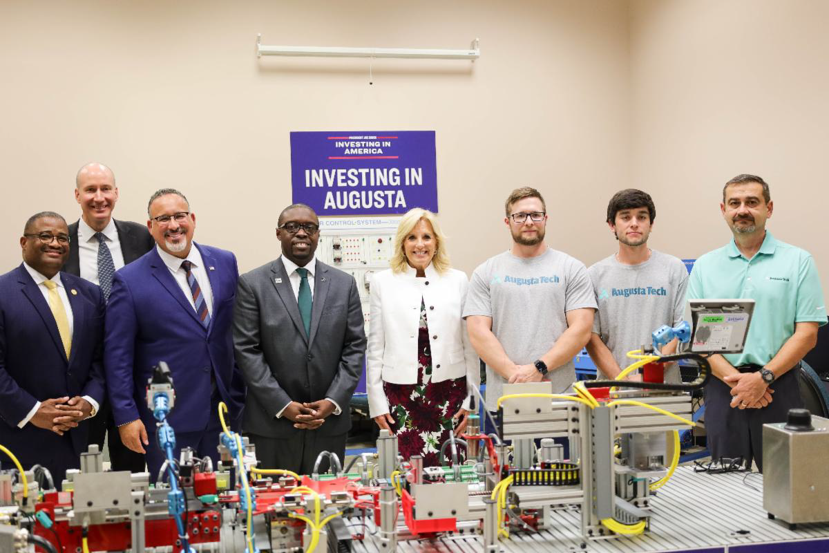 A photo of Mayor Garnett Johnson, Deputy Secretary of Energy David Turk, US Secretary of Education Dr. Miguel Cardona, President of Augusta Tech Dr. Jermaine Whirl, First Lady of the United States Dr. Jill Biden, student Dustin Phillips, student Patrick Kling and Mechatronics Instructor Ted Herlo standing in front of a mini assembly line in front of a blue sign with the words Investing in America and Investing in Augusta in white font on the wall.