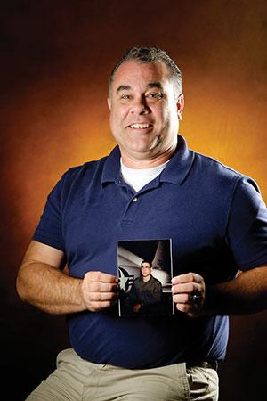 A middle-aged Caucasian male, Joey Turner is pictured smiling and wears a collared, short-sleeved, navy blue golf shirt with a white undershirt and holds a picture. He is standing against an orange background that fades to black around the edges.