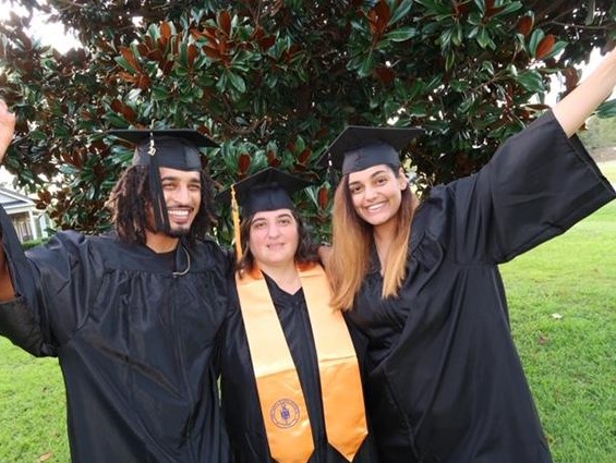 Smiling African American male with dreadlocks wearing a black graduation gown, black graduation cap with gold tassel, gold necklace and a blacks shirt; hand is spread out; center: Caucasian female with brown hair wearing a black graduation gown, black graduation cap with a gold tassel and a gold stole with a logo, left: smiling, female with golden brown hair wearing a black graduation cap, black graduation gown; hand is spread out; group is standing in front of a tree