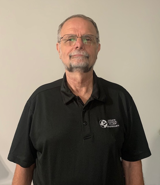 Caucasian presenting male wearing glasses with facial hair, wearing a black collared short sleeved shirt with a white Cybersecurity logo standing behind a plain background.