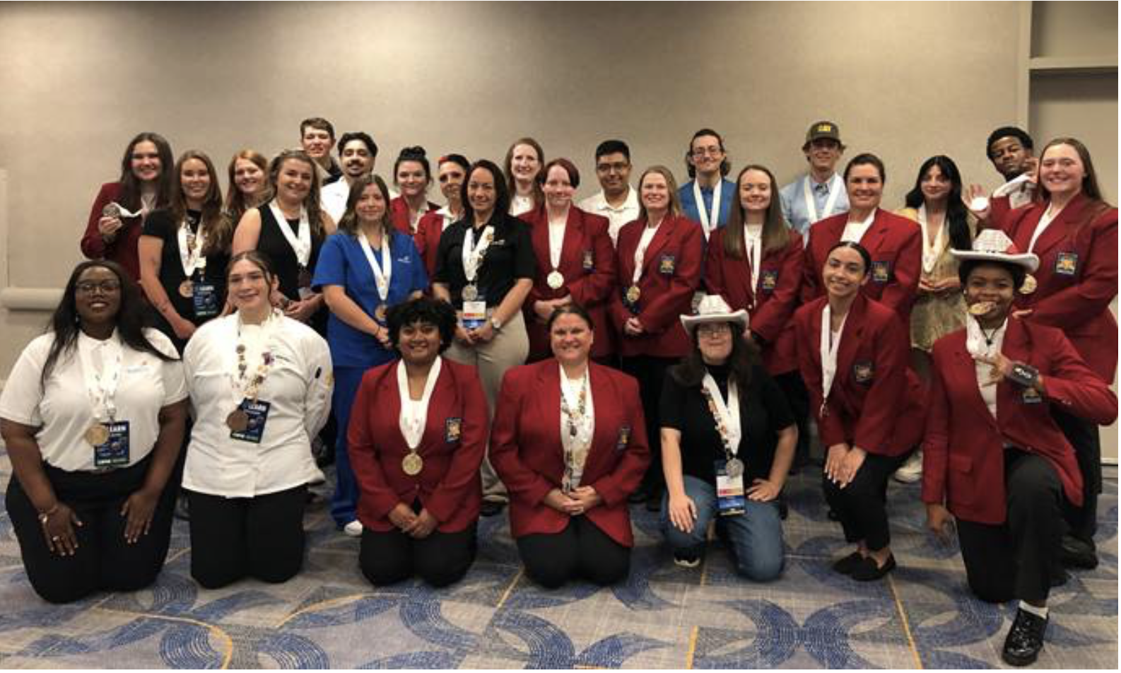 Group photo of approximately 30 SkillsUSA students and advisors posing together after an awards ceremony. Many participants wear red SkillsUSA blazers and medals around their necks, while others wear professional attire. The group is arranged in three rows inside a conference room, smiling toward the camera.
