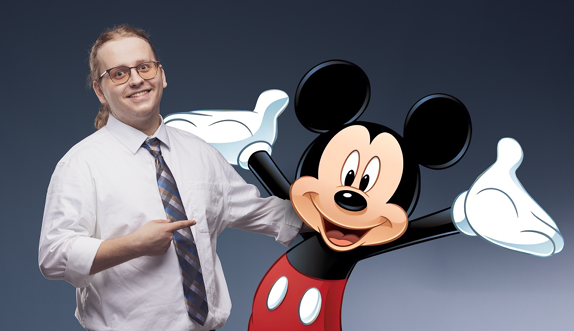 A smiling man in a white shirt and tie points toward Mickey Mouse standing beside him.