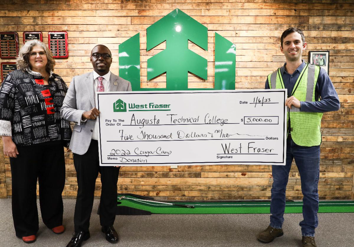 Rebecca Stout, Dr. Jermaine Whirl, and West Fraser General Manager Alex Moses stand holding the donation check in front of the west fraser logo on a wall composed of rustic, wooden, horizontal boards.