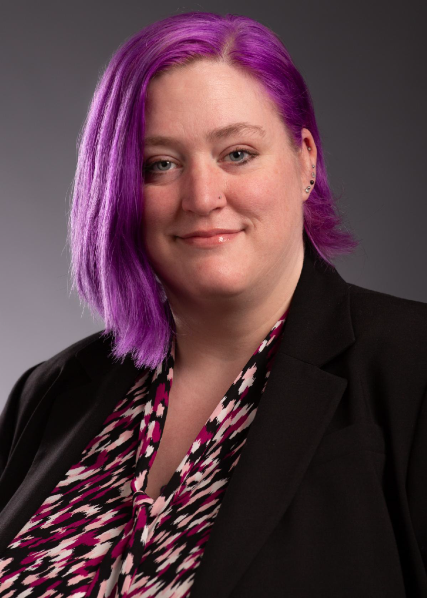 Ms. Alina Walls, a Caucasian female, smiles at the camera with chin length lavender hair wearing a red black and white patterned dress shirt with a black dress jacket against a grey background.