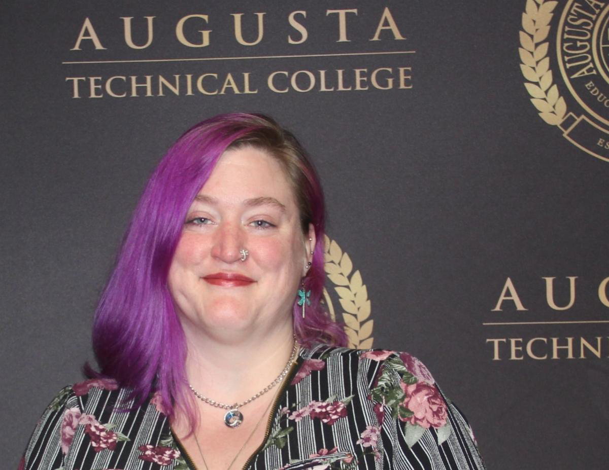 Alina Walls, a caucasian female with lavendar hair stands in front of a background with the ATC seal and name in gold and black. She is pictured wearing a black and gold patterned top.