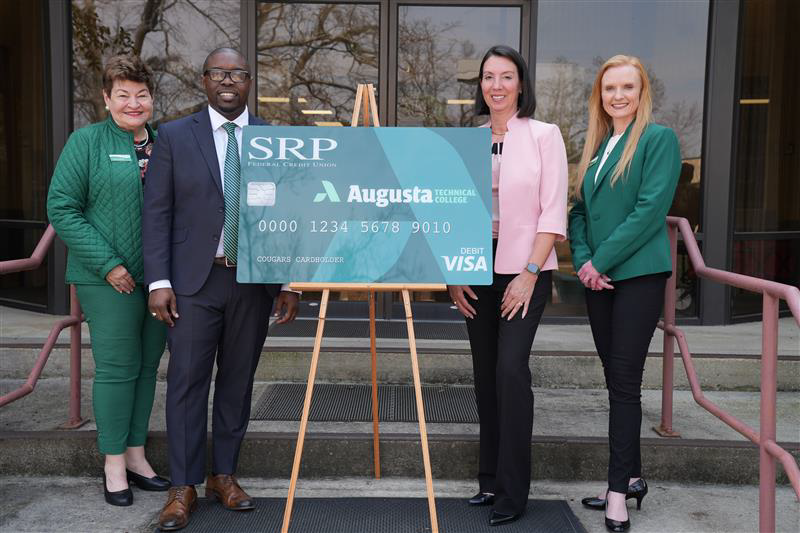 Ms. Yvette Meeks, Dr. Jermaine Whirl, Mrs. Shana Reid, and Ms. Grace Helms stand around a poster of the new debit card on the steps of the 100 building.