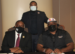 Three African American males wearing face masks and business attire posing for a photo; two males are sitting, one male is standing, both males who are sitting have their hands clasped in front of them on a table.