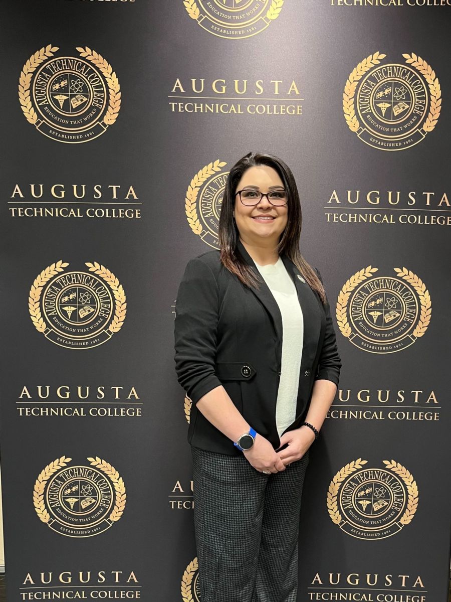 Devlynn Taylor, a caucasian female, stands in front of a backdrop with the Augusta Technical College seal and name in black and gold. She is wearing a black blazer with a white crew neck dress shirt and dark grey dress pants.