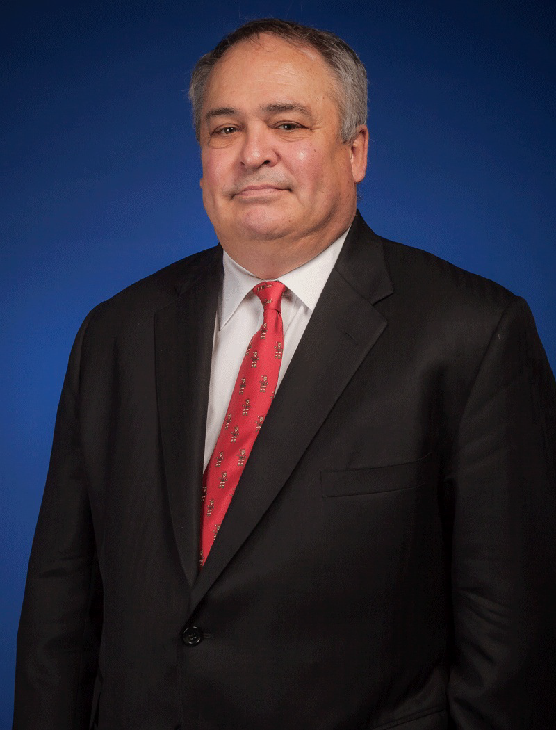 Smiling Caucasian male wearing a black suit, red tie, white collared shirt, posing for a photo against a dark dark blue background