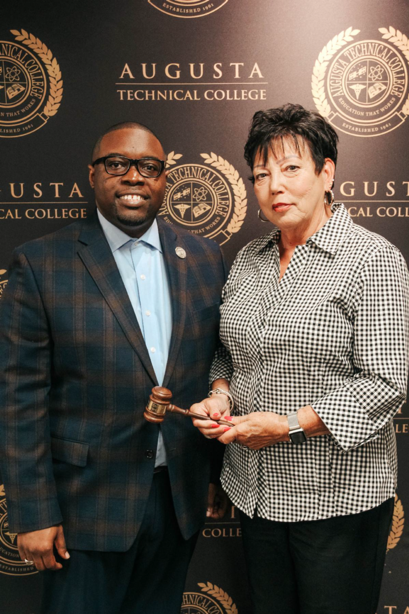 African American male wearing black glasses, blue collared shirt, blue blazer with a silver lapel pin and blue pants poses for a photo next to a Caucasian female wearing a black and white collared shirt and black pants and silver watch holding a brown gavel; both are standing in front of a black background with the repeating pattern of the Office of the President seal.