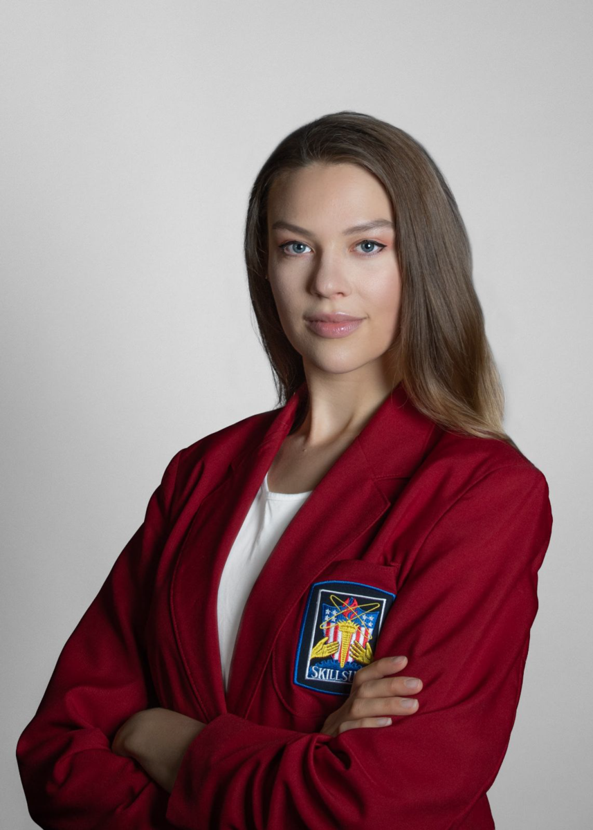Emily Pinto, a Caucasian female with long blonde hair, wears the red SkillsUSA jacket and a white shirt in front of a light grey background.