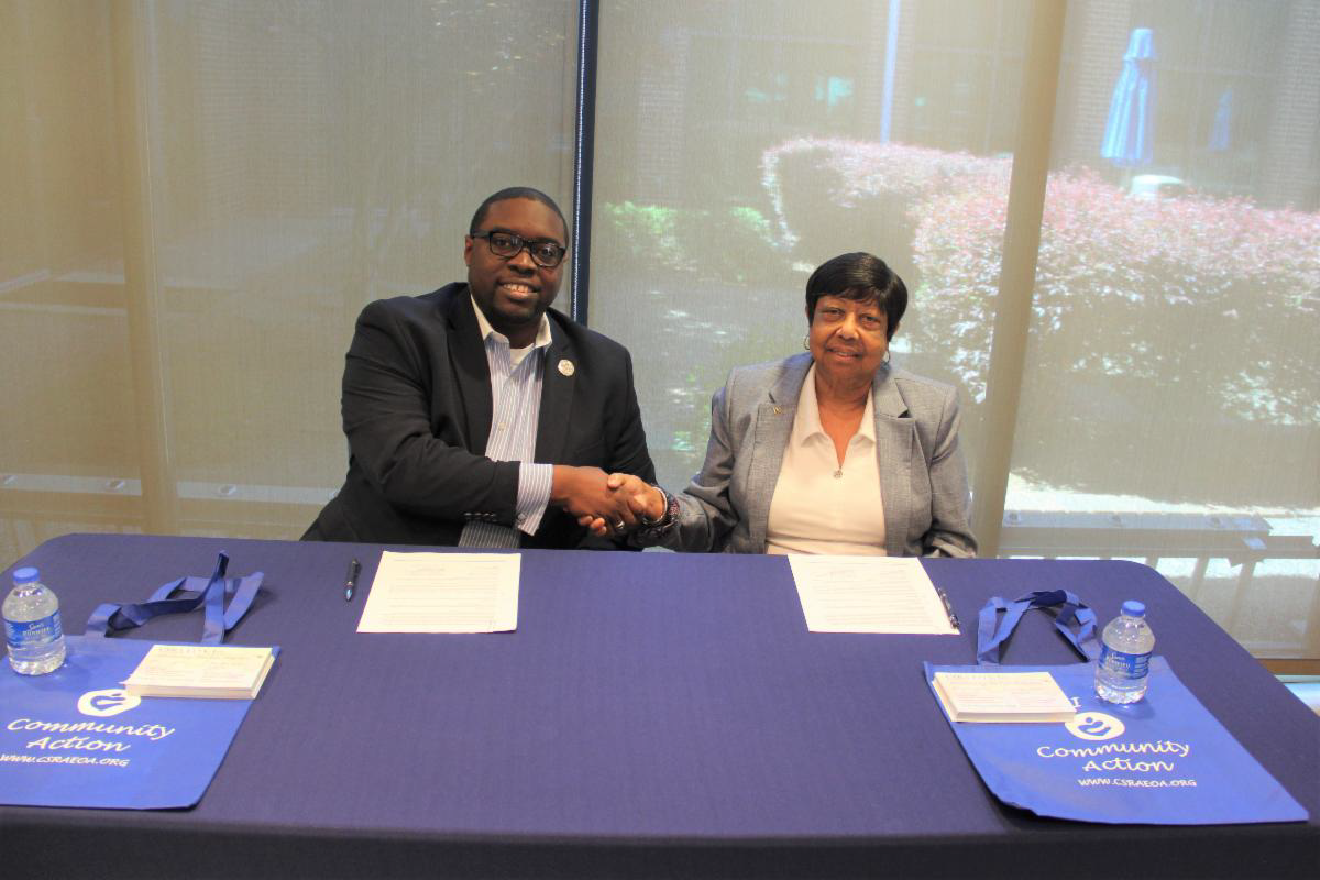 African American male wearing a blue blazer, white collared shirt, silver lapel pin, sitting in a chair shaking hands next to an African American female wearing a gray blazer, white shirt, both are sitting in front of a table with a blue table cloth, on the table are white papers and blue and white bags. The photo is in front of a window, and outside the window are flowers.