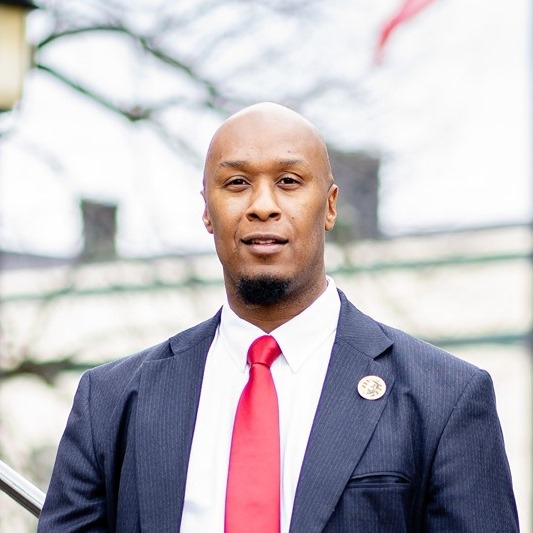 Judge Ashley Moore, an African American male, wears a blue suit with a white collared shirt and red tie while standing outdoors in an urban area.