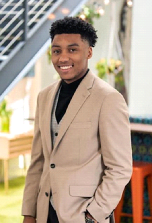 Smiling, young, African American male wearing a tan-colored business suit with black buttons and pockets, black shirt, and gray vest with his hand in his pocket; there is a  chair, desk, flowers, and stair case in the background.