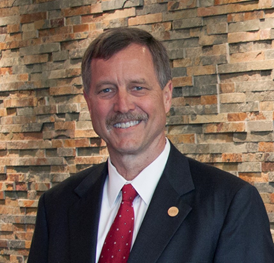 Smiling, Caucasian male with a mustache wearing a blue suit, white collared shirt, red tie with print, gold lapel pin, standing against a brick wall, posing for a photo