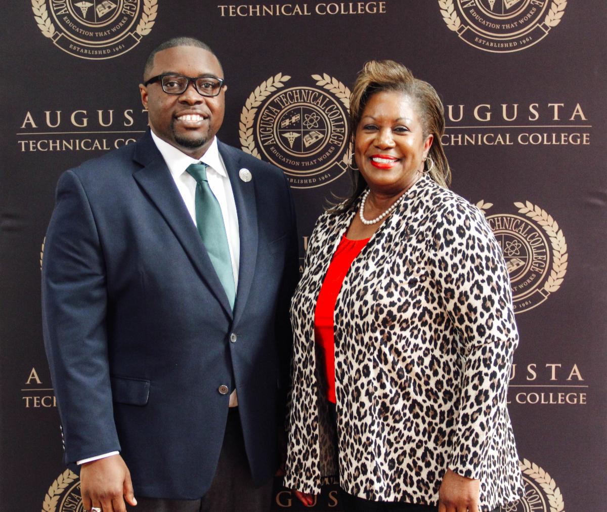 Photo: Smiling African American male wearing black glasses, blue suite, light blue/teal tie, white collared shirt, with silver lapel pin standing next to a smiling African American female wearing red shirt, pearl necklace, a leopard print cardigan, posing for a photo in front of a black background with the Augusta Tech seal.