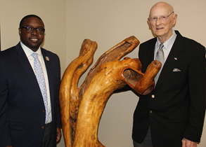 African American male, wearing black glasses, a blue suit, white collared shirt, light blue tie, silver Augusta Tech lapel pin, standing next to a wood statue; Caucasian male wearing glasses, a black suit, blue collared shirt, pocket square; both are posing for a photo against a tan-colored wall