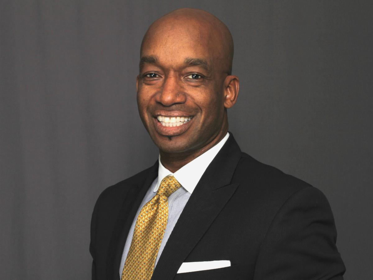 Smiling African American male wearing a black blazer, white collared shirt, yellow tie with black dots and a white napkin in his upper coat pocket posing for a photo