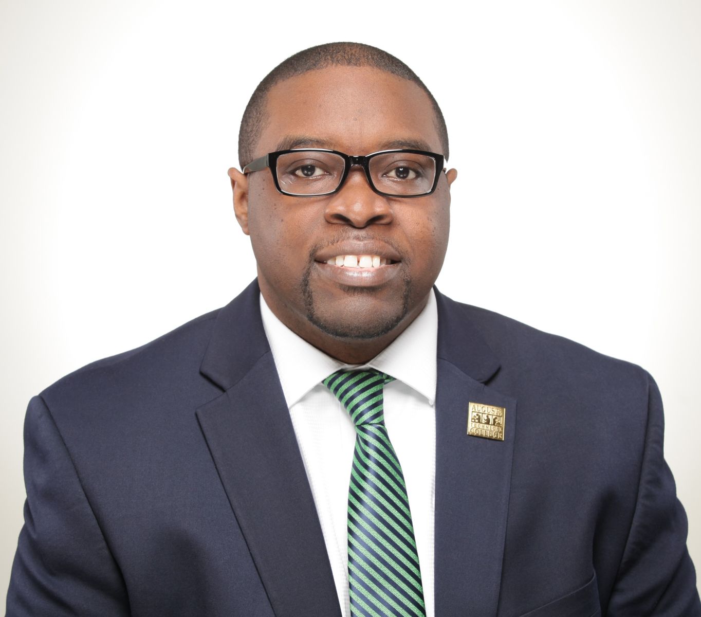 Smiling, African American male wearing black glasses wearing a blue suit, white collared shirt, green tie with blue stripes, and gold Augusta Technical College lapel pin.